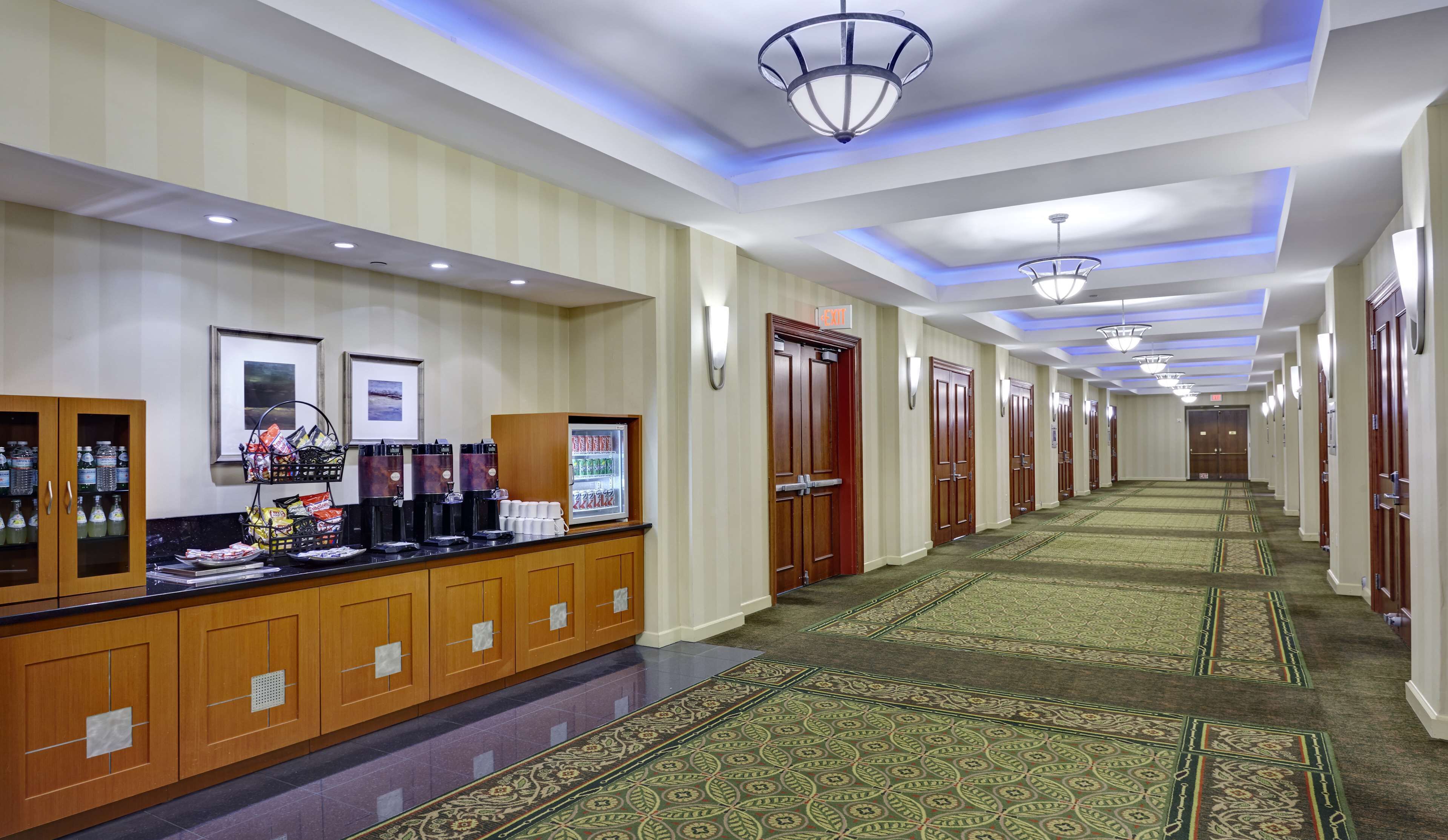 A hotel corridor with a reception area featuring a coffee station, framed pictures, and a carpeted floor.