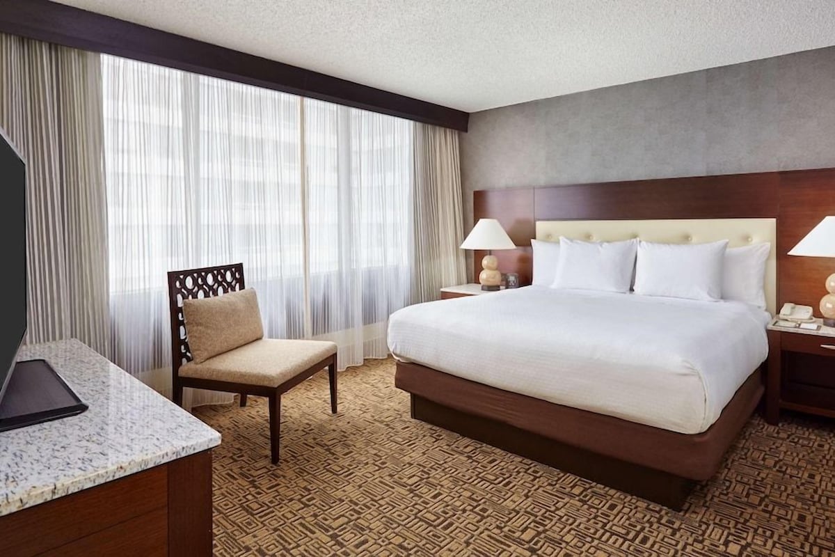 A hotel room with a large bed, a chair, a desk with a television, and a window with sheer curtains.