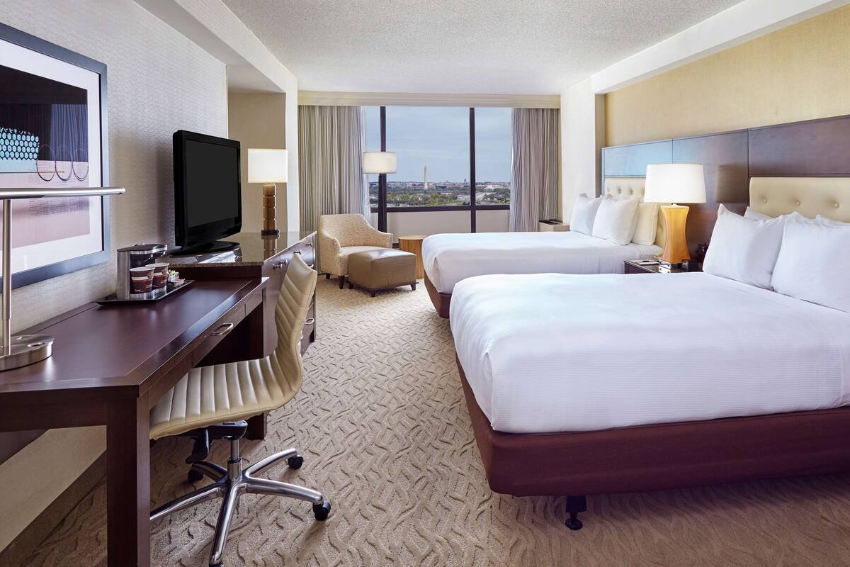 A hotel room with a desk, two beds, a TV, and a view of the city.