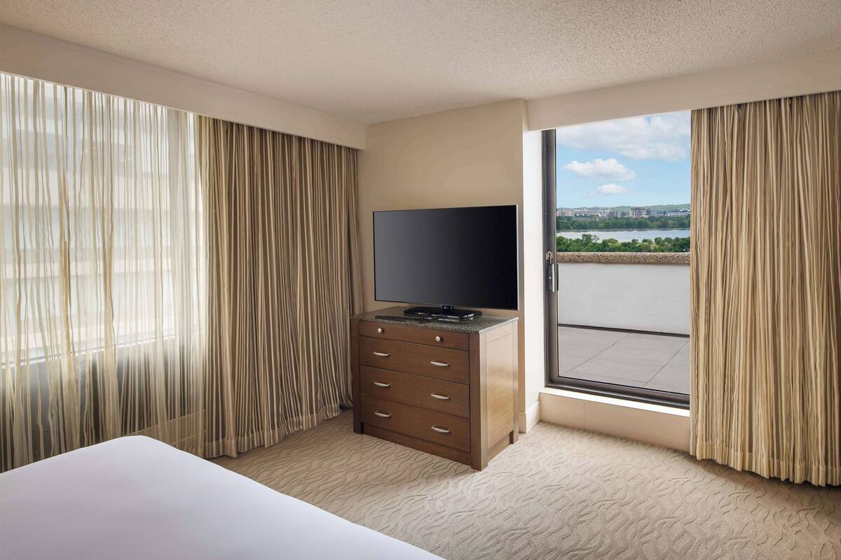 A hotel room with a bed, a TV on a wooden cabinet, and a view of a body of water through a large window.