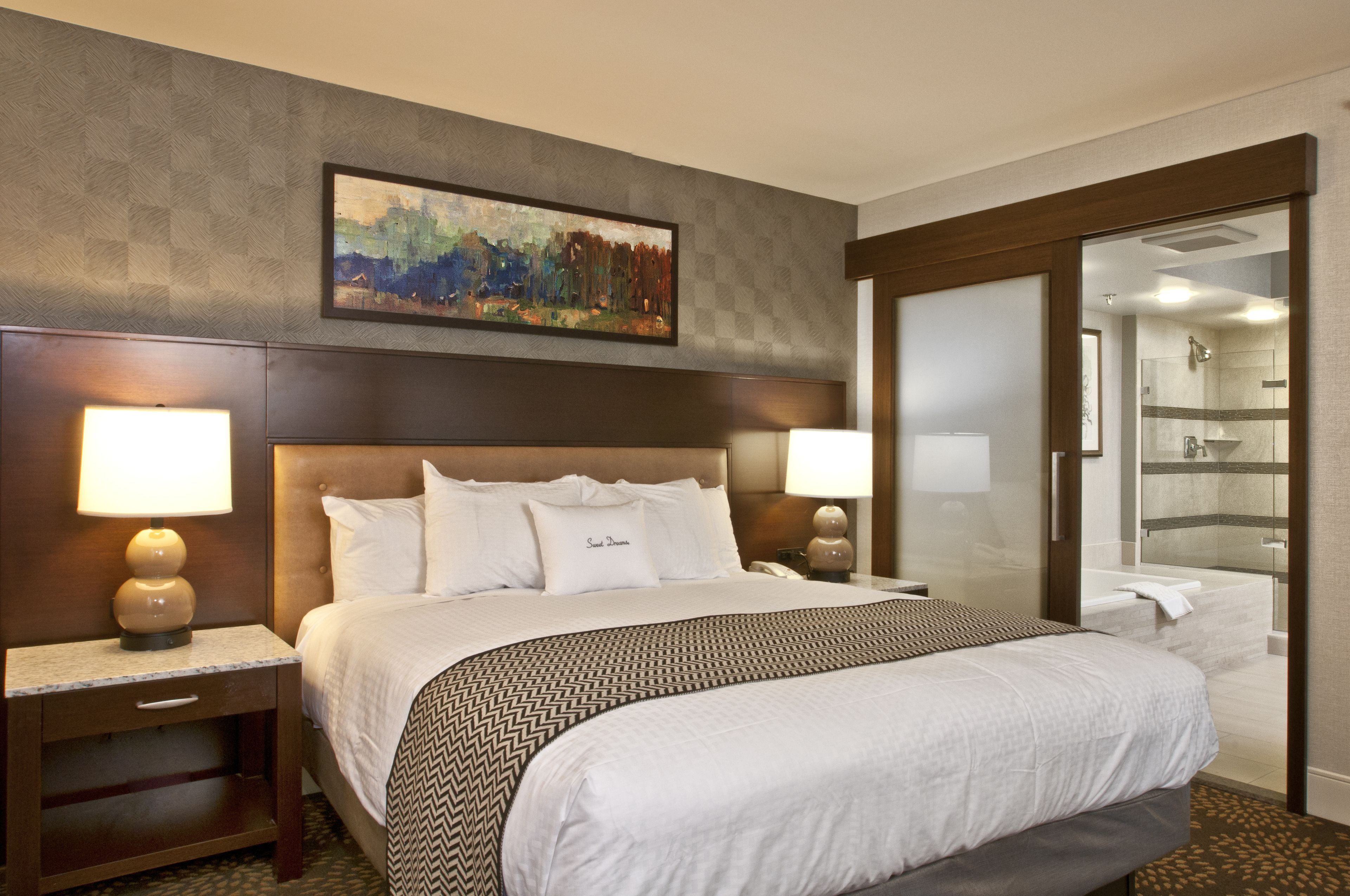 A hotel room with a large bed, two bedside tables with lamps, a painting on the wall, and a bathroom visible through an open door.