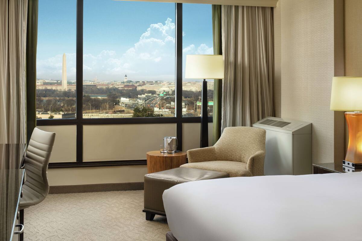 A hotel room with a large window offering a view of the Washington Monument, a bed, a chair, a side table with a lamp, and a heater.