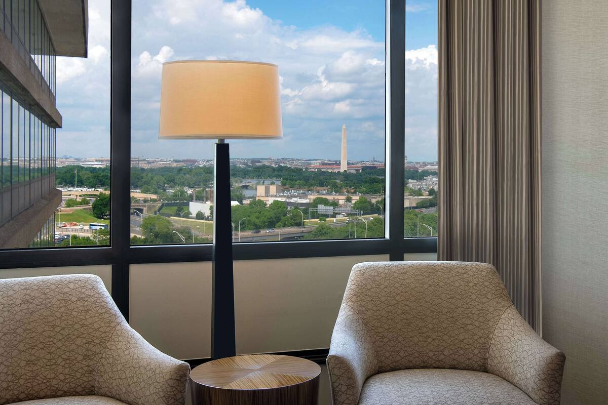 A hotel room with two armchairs, a small wooden table, and a view of the Washington Monument through large windows.