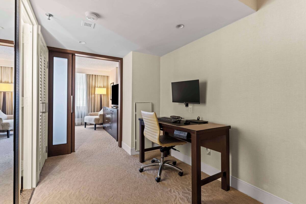 A hotel room with a desk, chair, and TV. There is a door leading to another room with a bed and curtains.