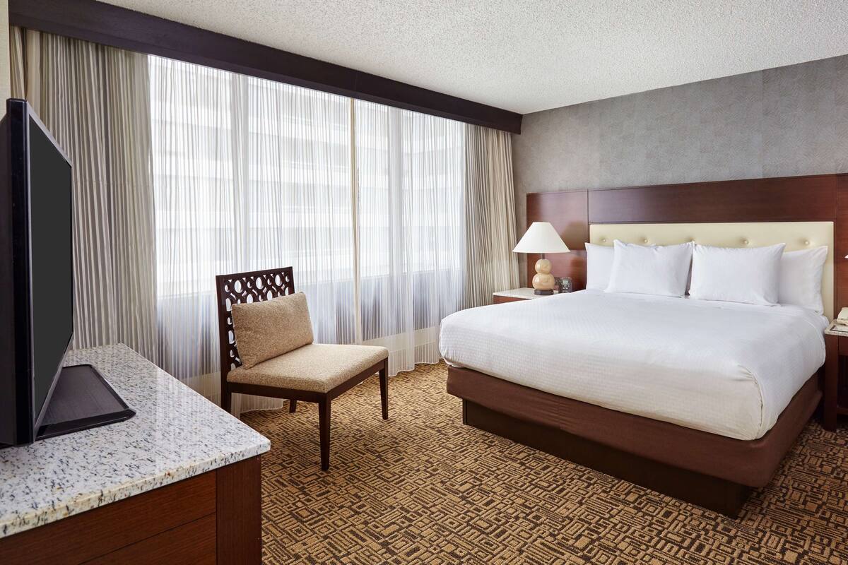 A hotel room with a bed, a chair, a desk, a TV, and a window with curtains.