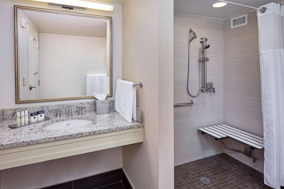 A bathroom with a marble countertop, two sinks, a large mirror, a towel rack, and a shower area with a bench.