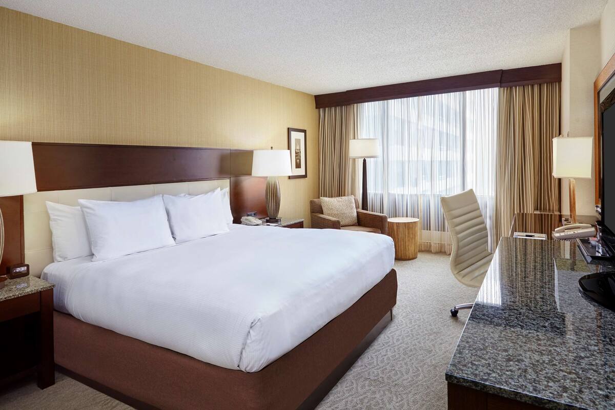 A hotel room with a large bed, a desk with a flat-screen TV, a chair, a lamp, and a bedside table with a phone.