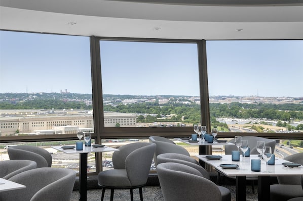 A restaurant with a view of a cityscape, featuring tables set with glasses and plates, and modern chairs.