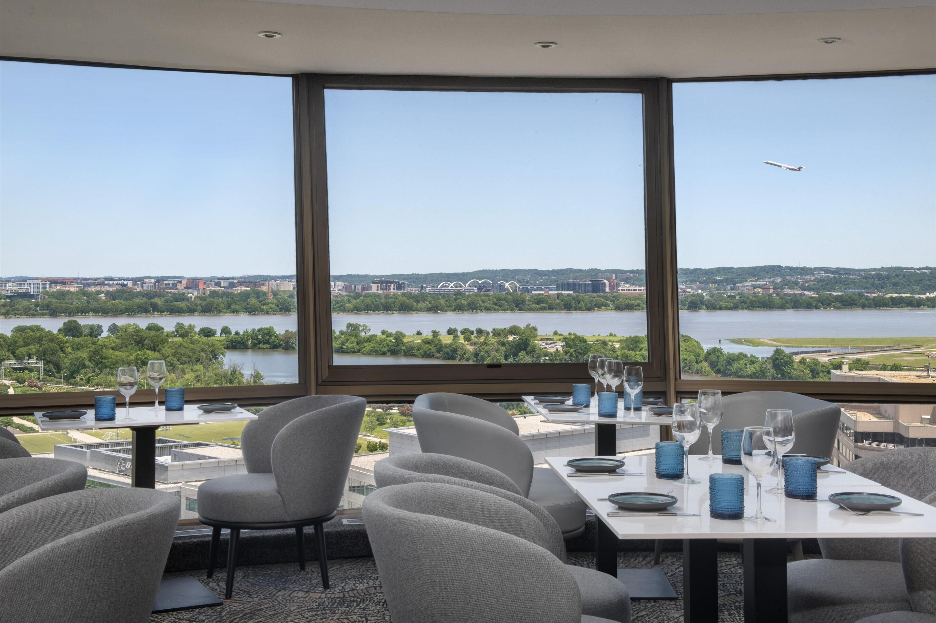 Ein Essbereich mit Tischen für zwei Personen, große Fenster mit Blick auf einen Fluss und eine Stadtansicht, sowie ein entfernt fliegendes Flugzeug.
