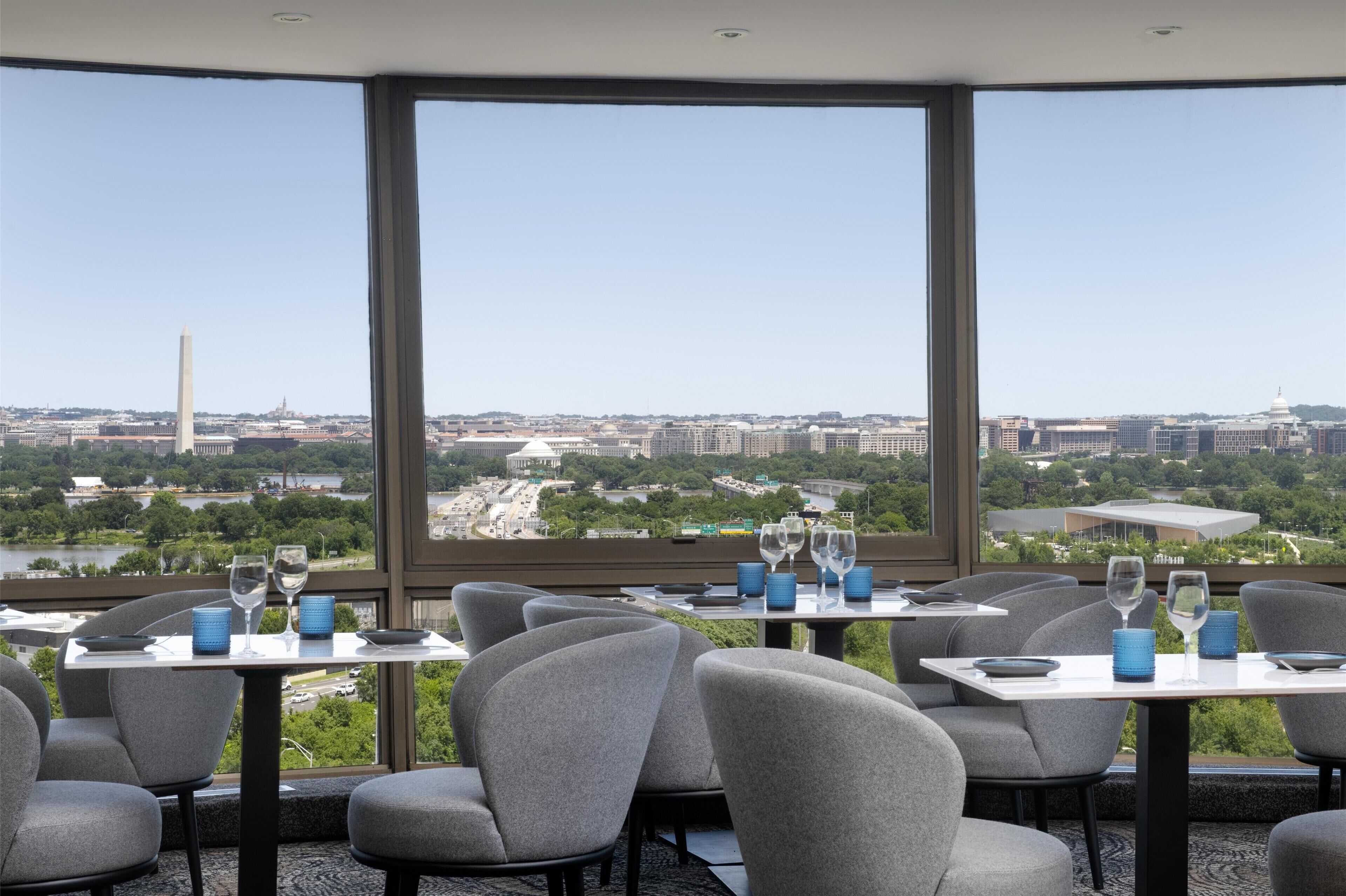 A restaurant with tables set for dining, large windows offering a cityscape view, and a clear sky.