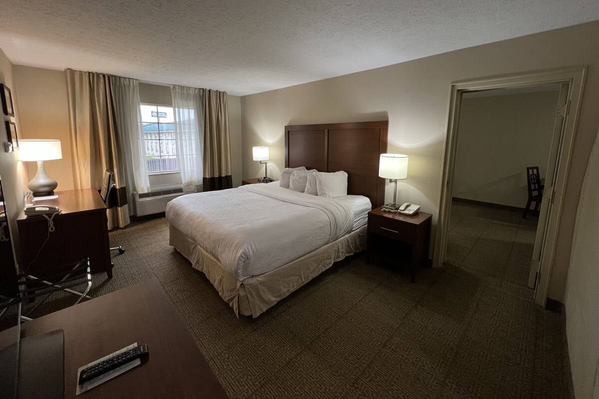 Een hotelkamer met een groot bed, twee nachtkastjes met lampen, een bureau met stoel, een raam met gordijnen en een deur naar een andere kamer.