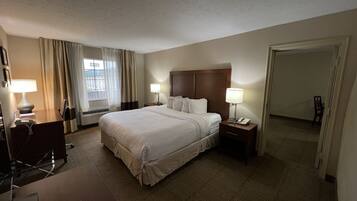 A hotel room with a large bed, two bedside tables with lamps, a desk with a chair, a window with curtains, and a door leading to another room.
