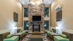 A lobby with a stone fireplace, green upholstered seating, and a large TV screen. - Wyndham Garden Cross Lanes Charleston (Cross Lanes)