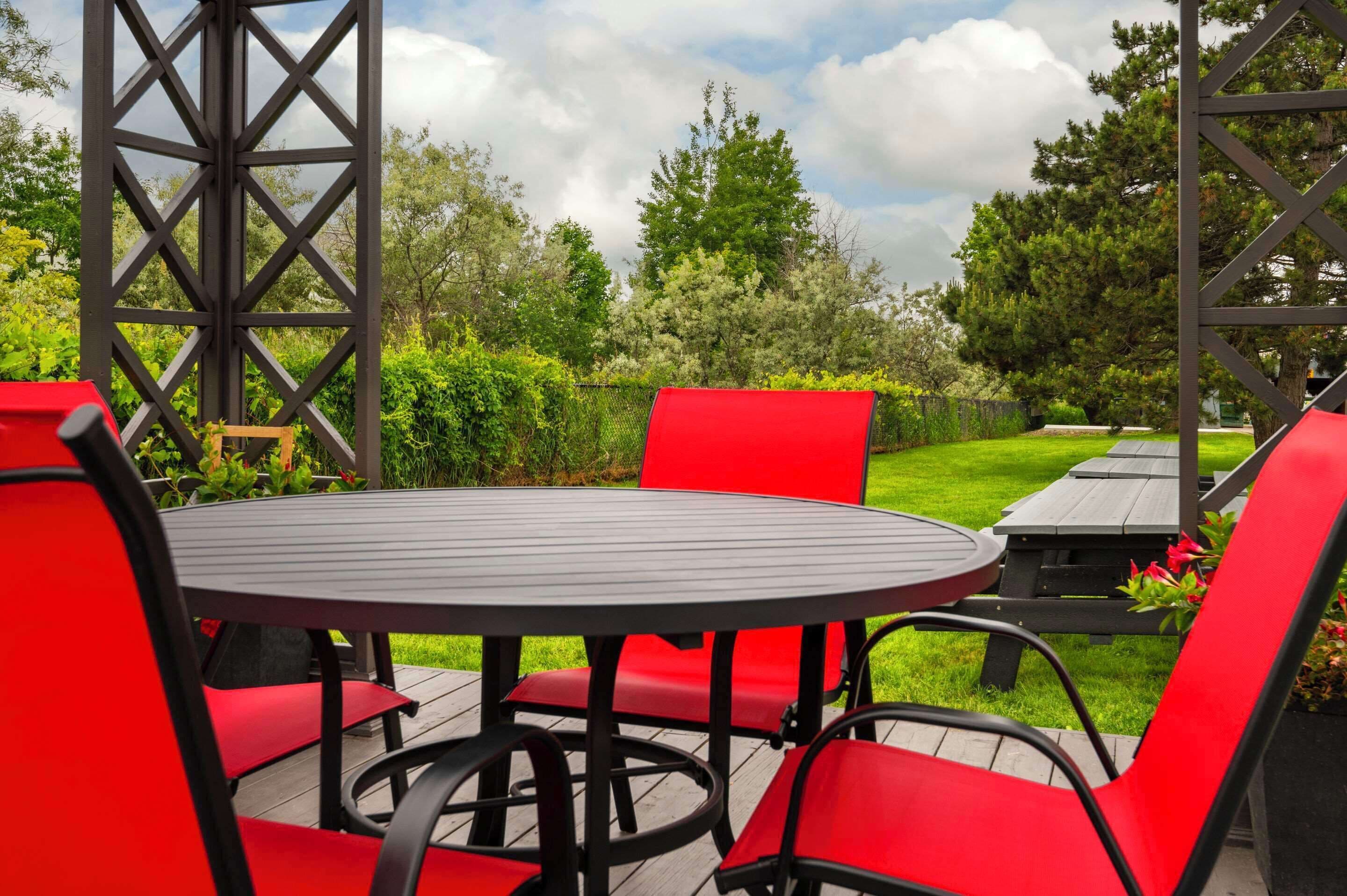 Une table ronde, dotée de chaises rouges et d’un cadre en métal noir, est placée sur une terrasse en bois, offrant une vue sur une zone herbeuse et des arbres en arrière-plan.
