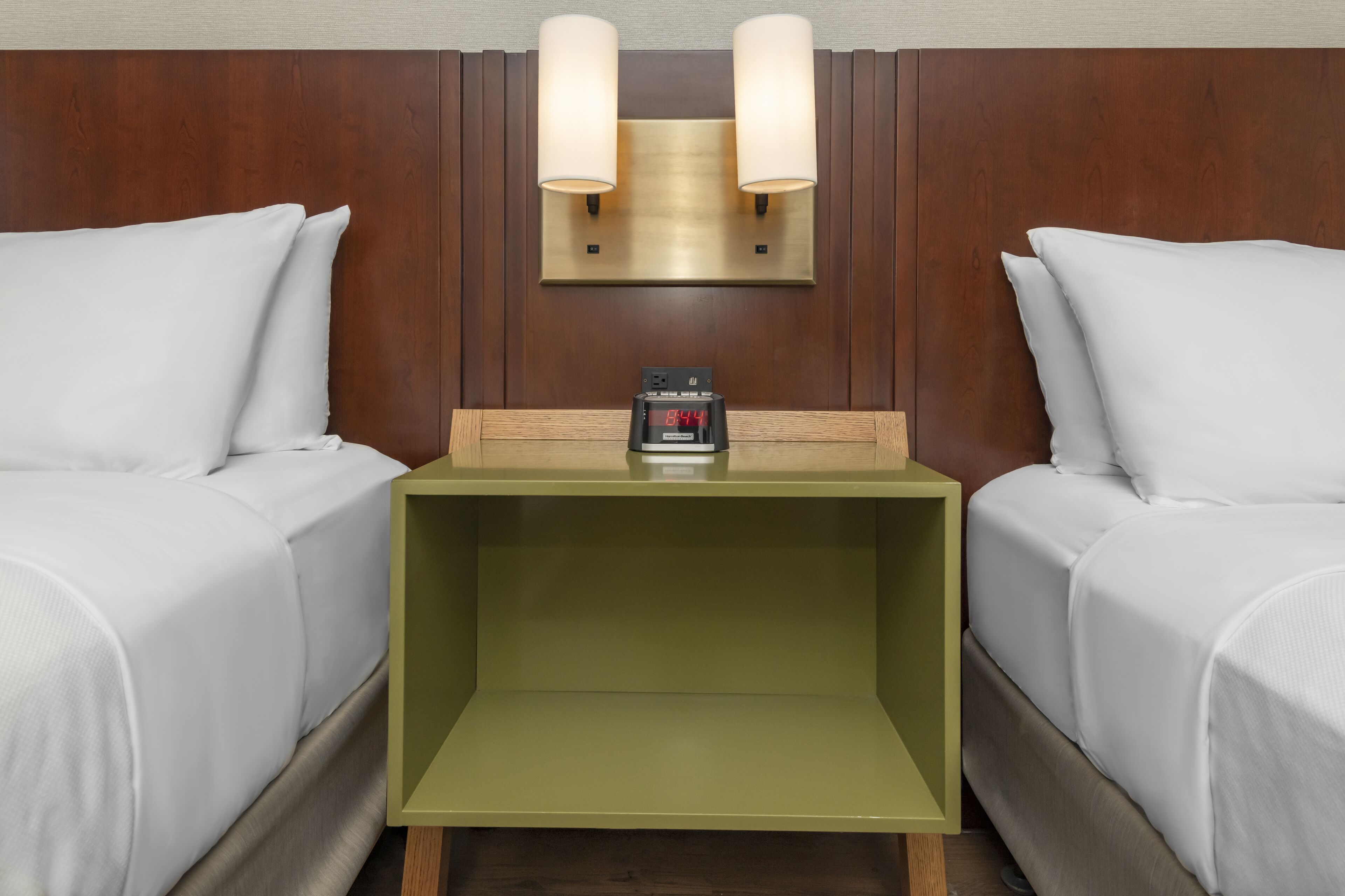 Una habitación de hotel con dos camas, una mesita de noche verde, un reloj digital e iluminación en la pared.