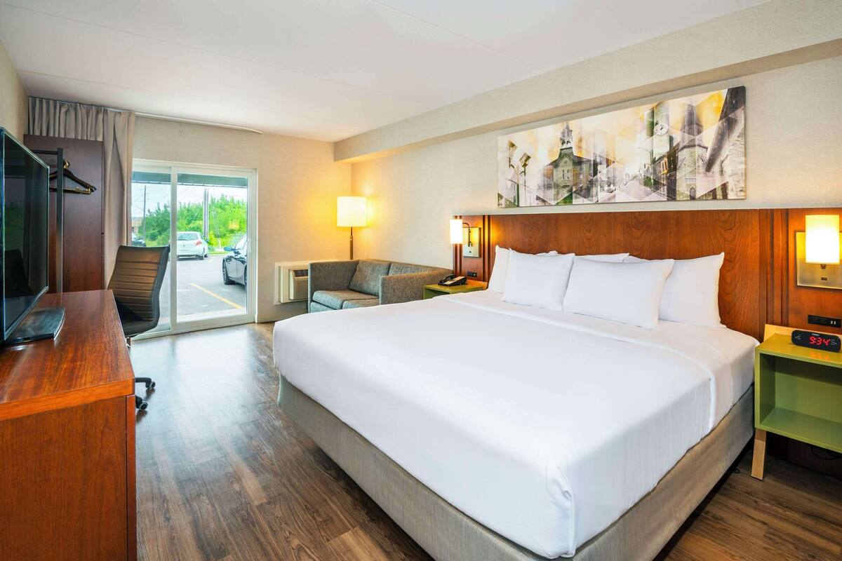 A hotel room with a large bed, a desk, a chair, a television, a painting on the wall, and a view of the outside.