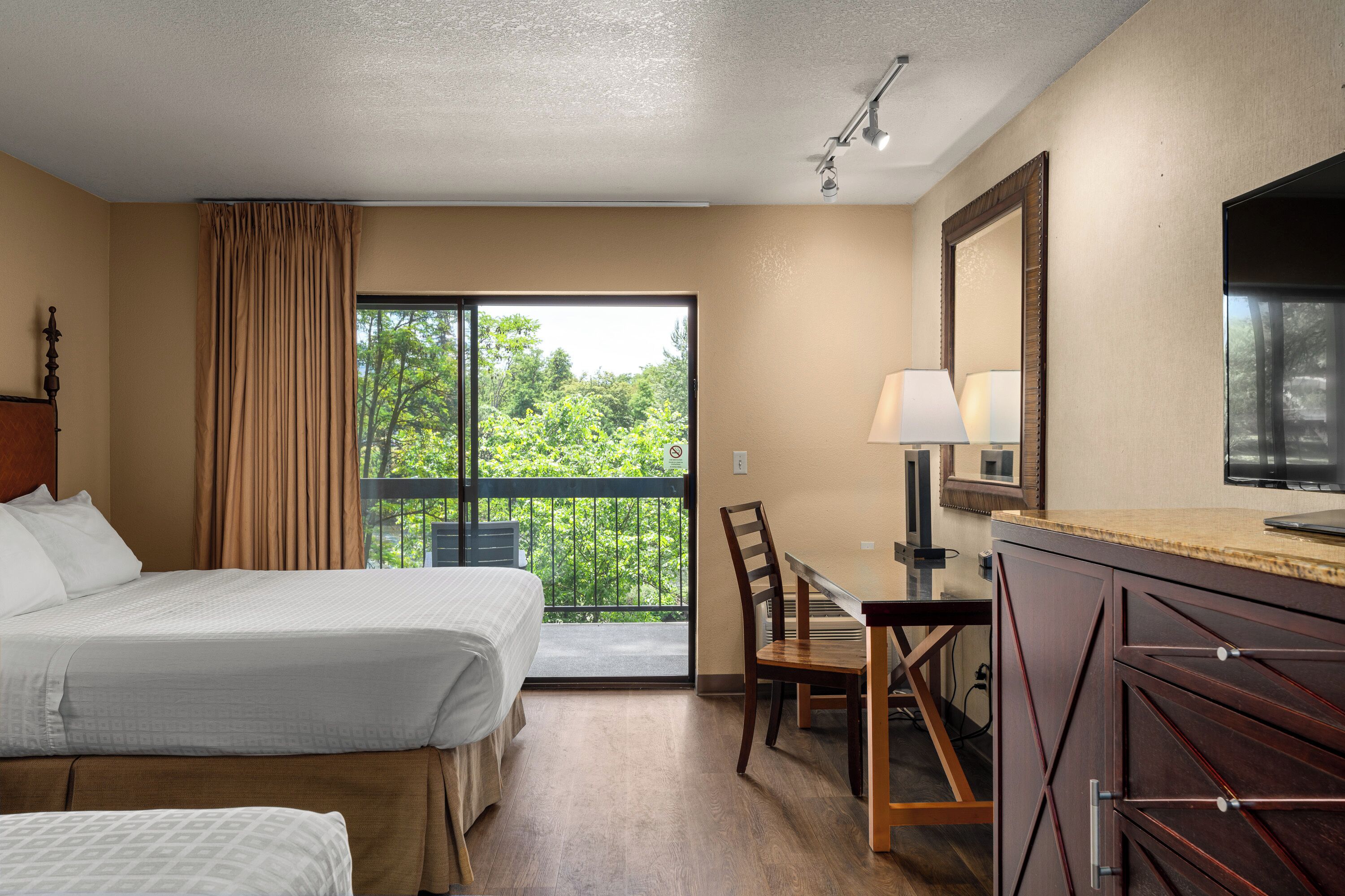 Hotelkamer met een bed, een bureau, een stoel, een televisie en een balkon met uitzicht op bomen.
