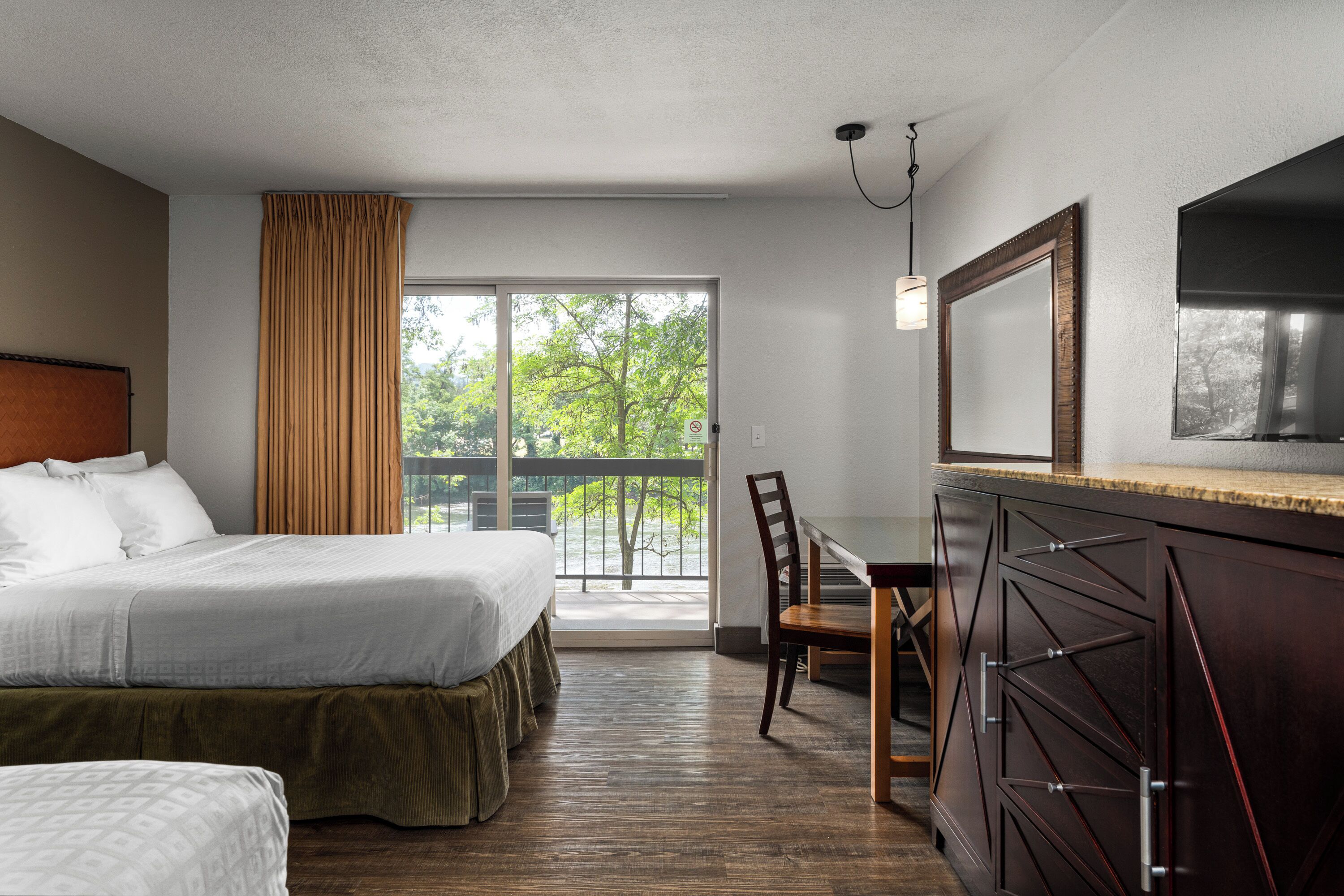 Ein Hotelzimmer mit Bett, Schreibtisch, Stuhl, Kommode und einem großen Fenster mit Blick auf Bäume.