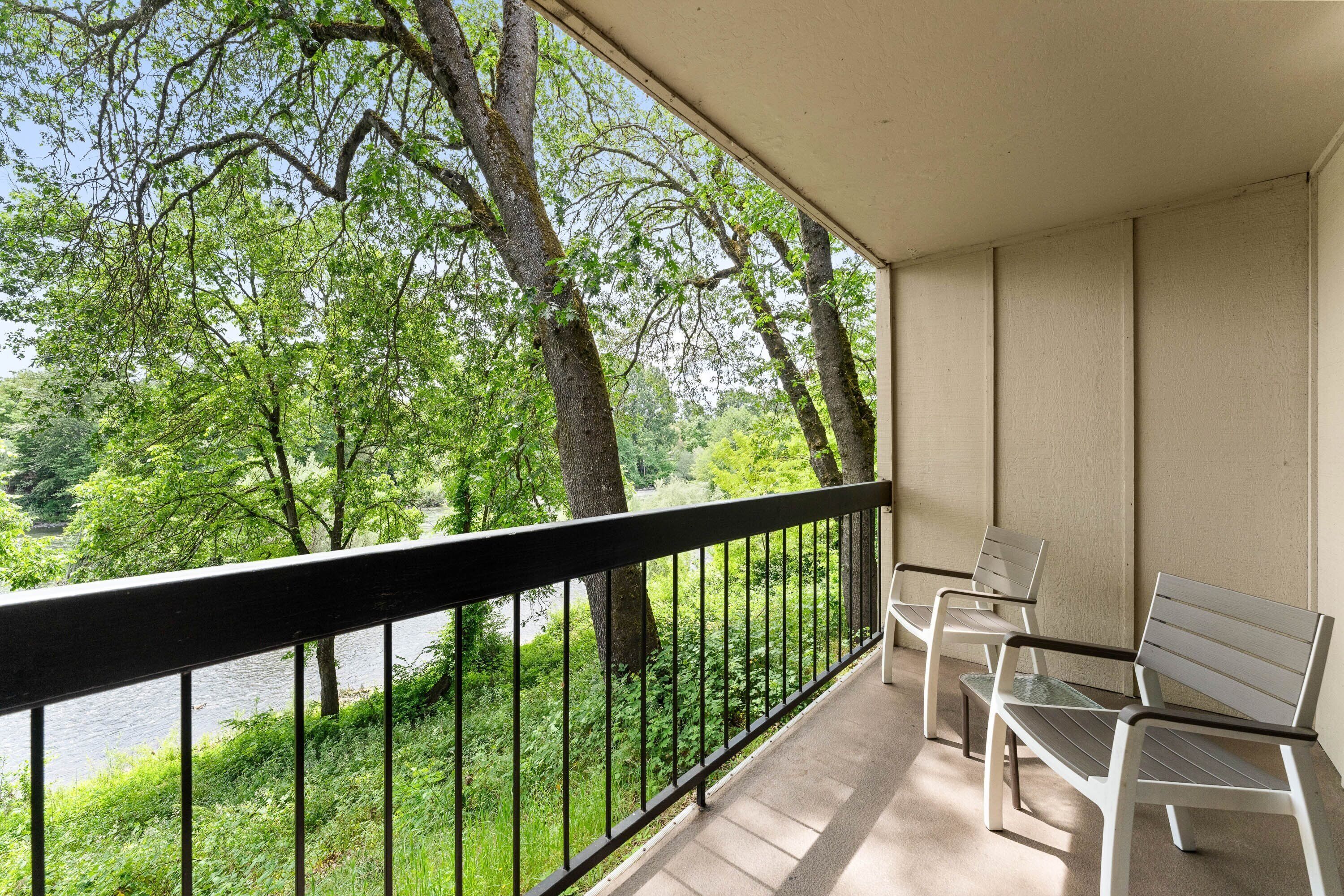 A balcony with a view of a river and trees, two white chairs, and a black railing.