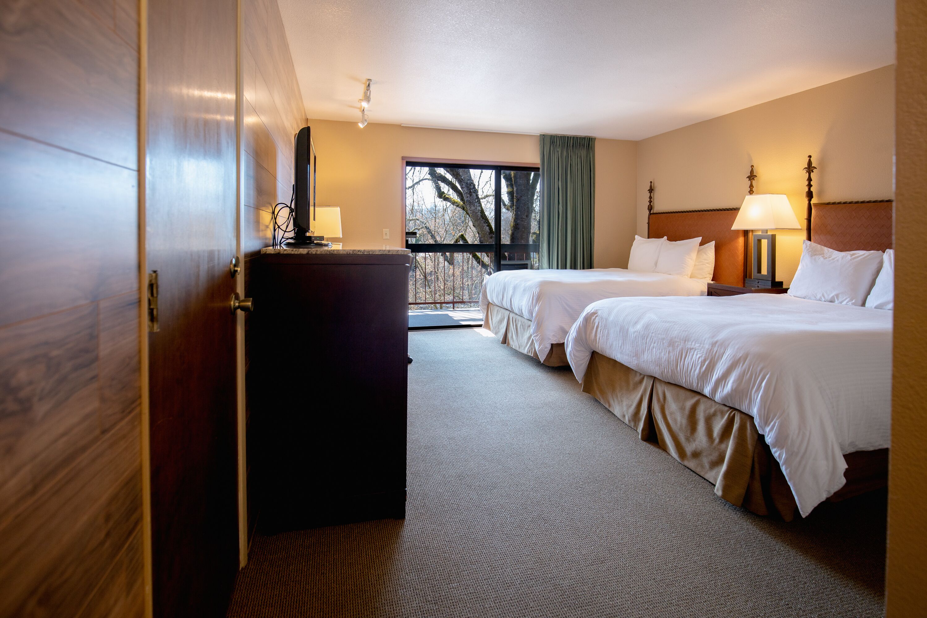 A hotel room with two beds, a desk, and a window with a view of trees.