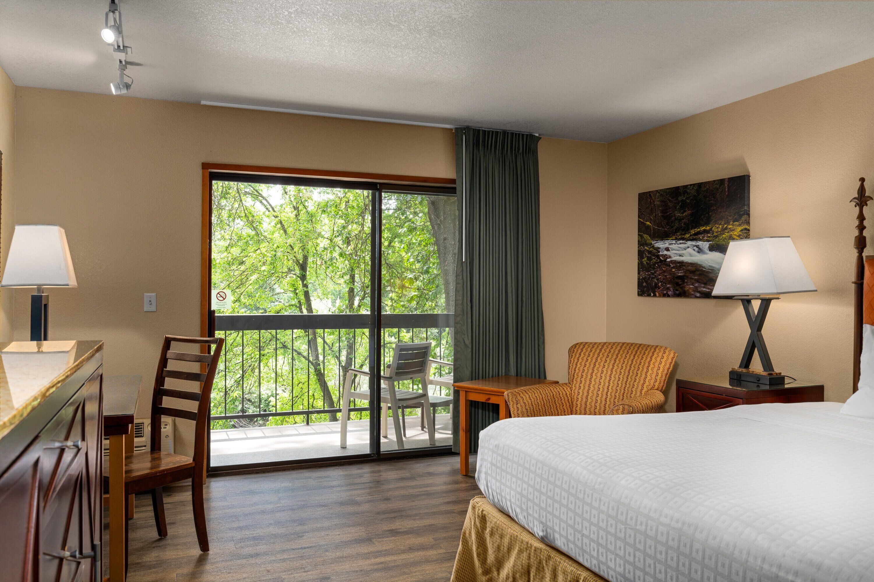 A hotel room with a bed, a desk, a chair, a table, a lamp, a painting, and a balcony with a view of trees.