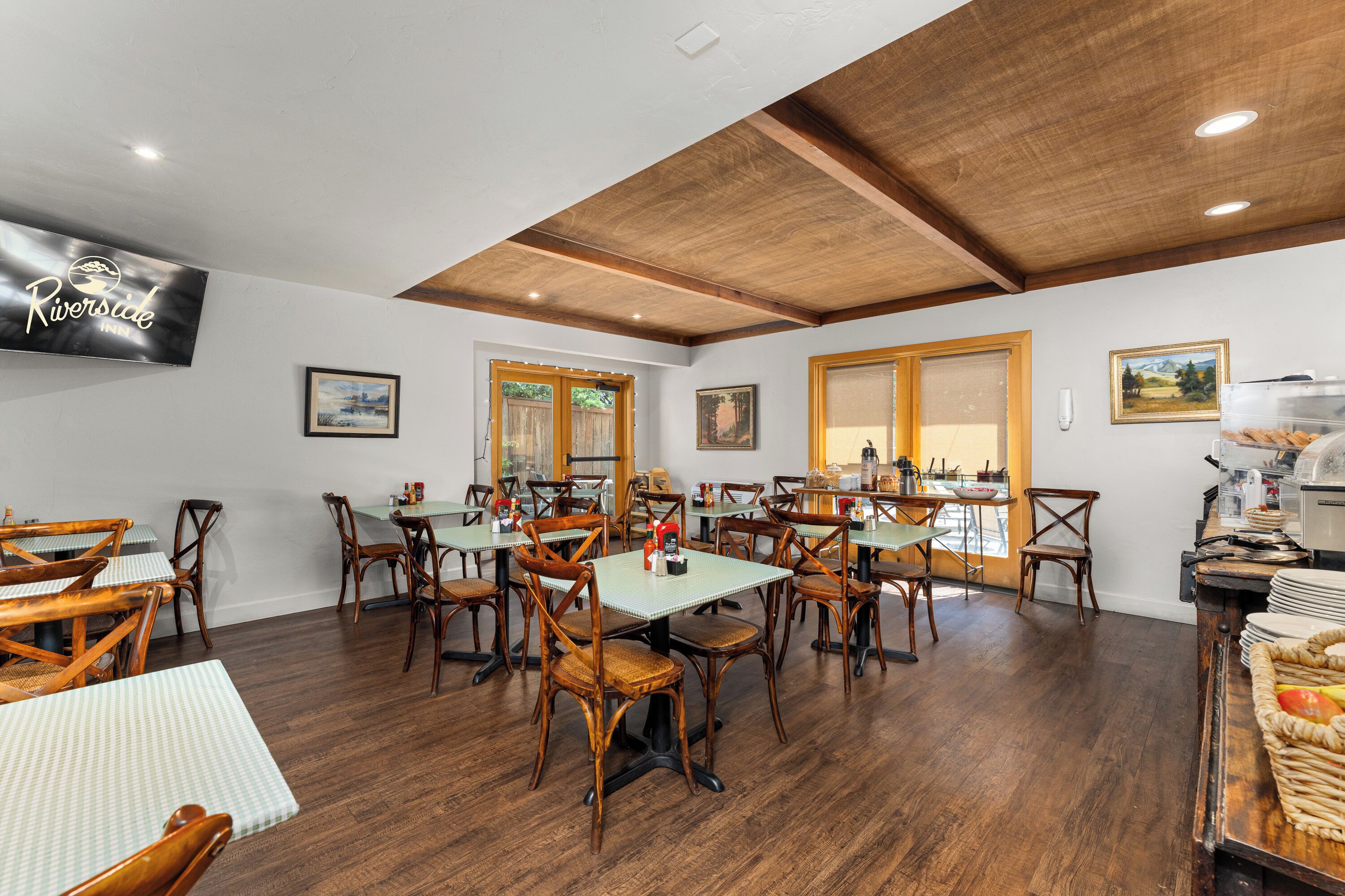 A dining area with wooden tables and chairs, a television displaying the Riversdale Inn logo, and framed artwork on the walls.