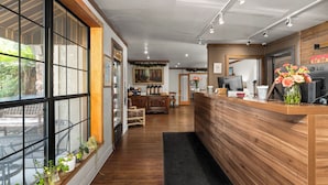 Zona de recepción con mostrador de madera, un ventanal con plantas, comedor con sillas y un cuadro en la pared.