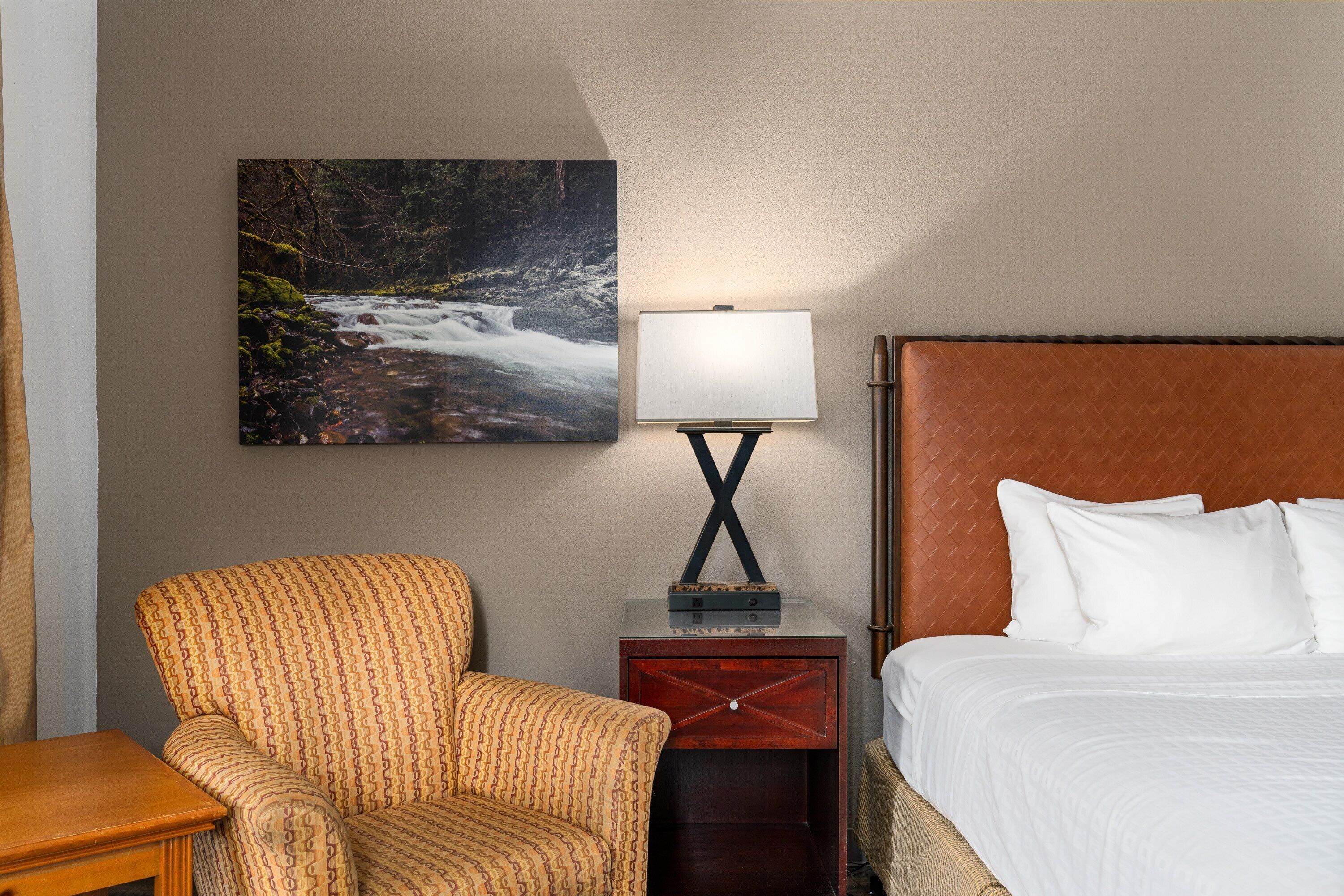Une chambre d’hôtel avec un lit, une chaise, une table de chevet avec une lampe et un tableau représentant une cascade.