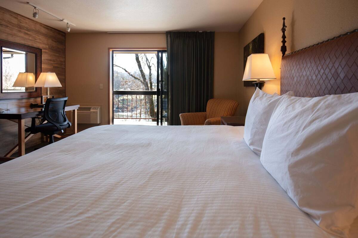 A hotel room with a large bed, a desk with a chair, a window with a view, and a lamp.