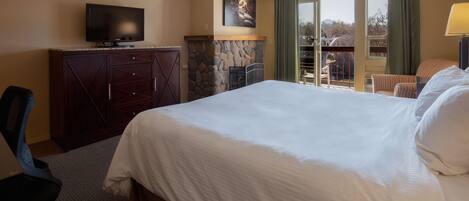 A hotel room with a large bed, a television on a wooden cabinet, a stone fireplace, a balcony with a chair, and a view of the outdoors.