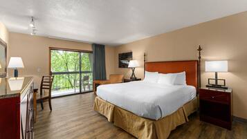 A hotel room with a large bed, a desk, a chair, a nightstand, and a window with a view of greenery.