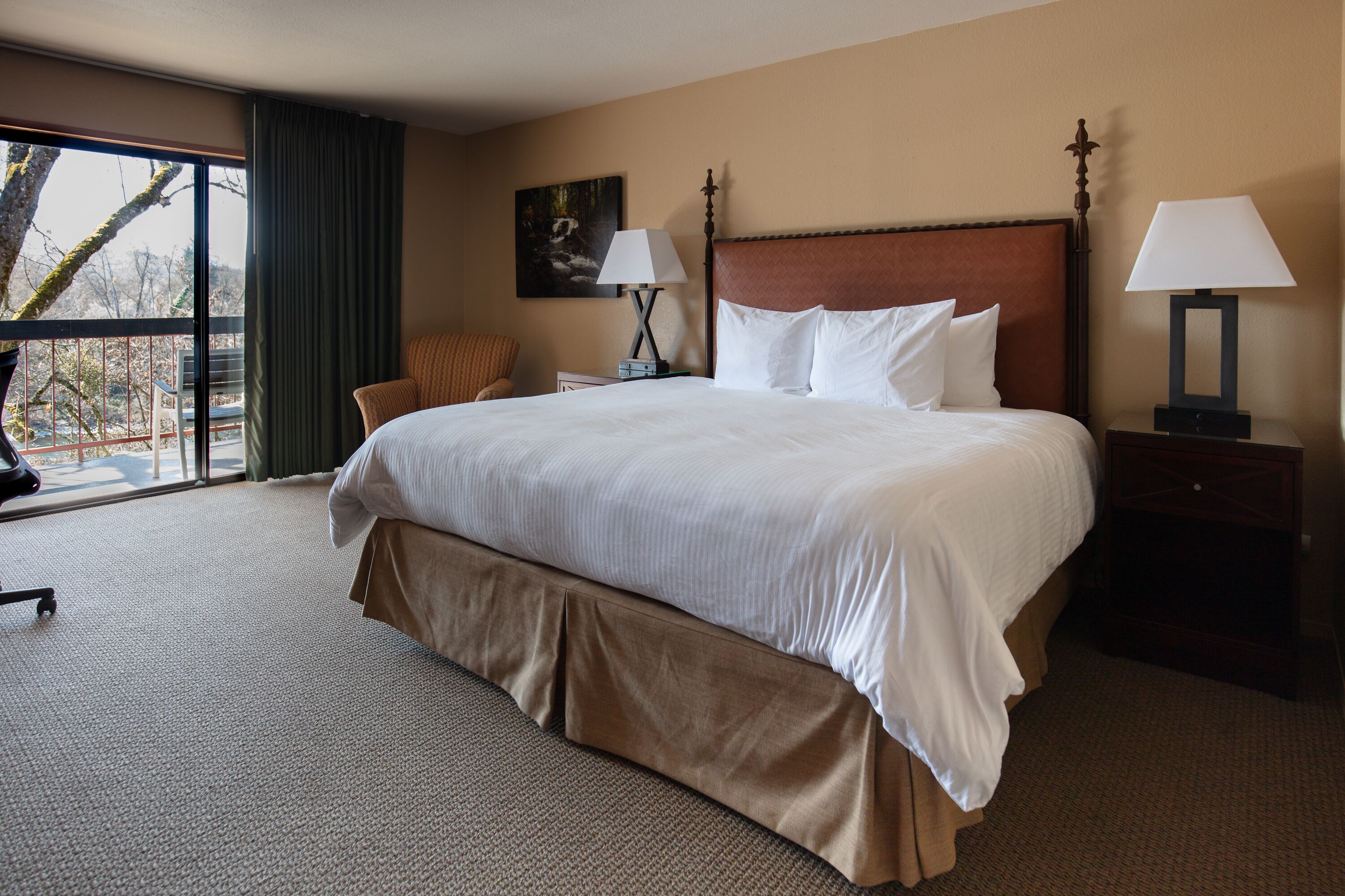 A hotel room with a large bed, two bedside tables with lamps, a chair, a painting on the wall, and a view through a window.