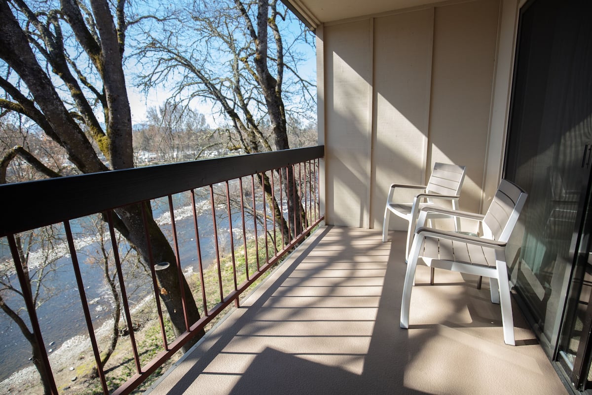 A balcony with a view of a river and trees, and two white chairs.