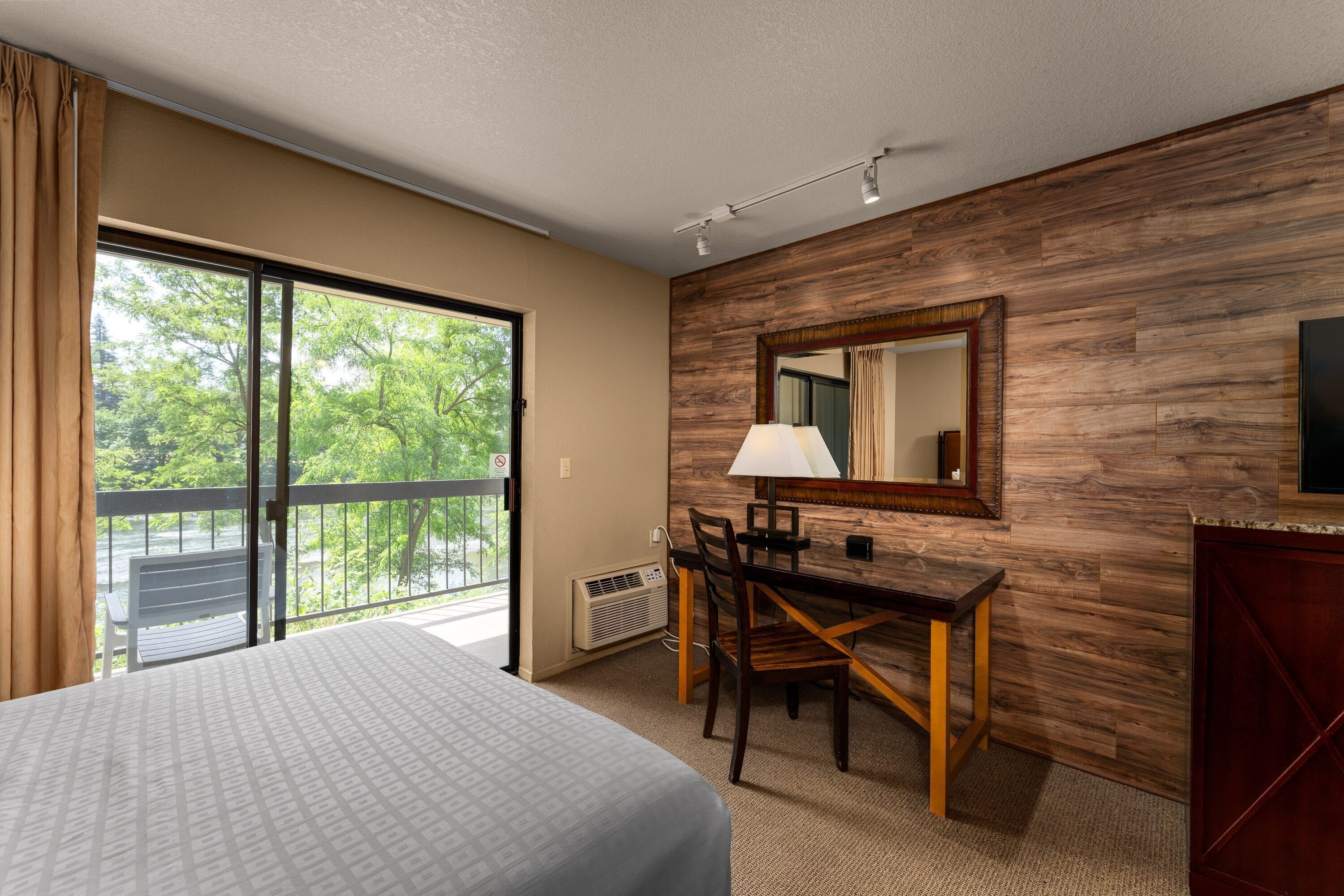 A hotel room with a bed, a desk, a chair, a lamp, a mirror, and a view of greenery outside.