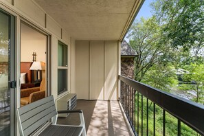A balcony with a wooden bench, a table with a lamp, and a view of greenery and a river.