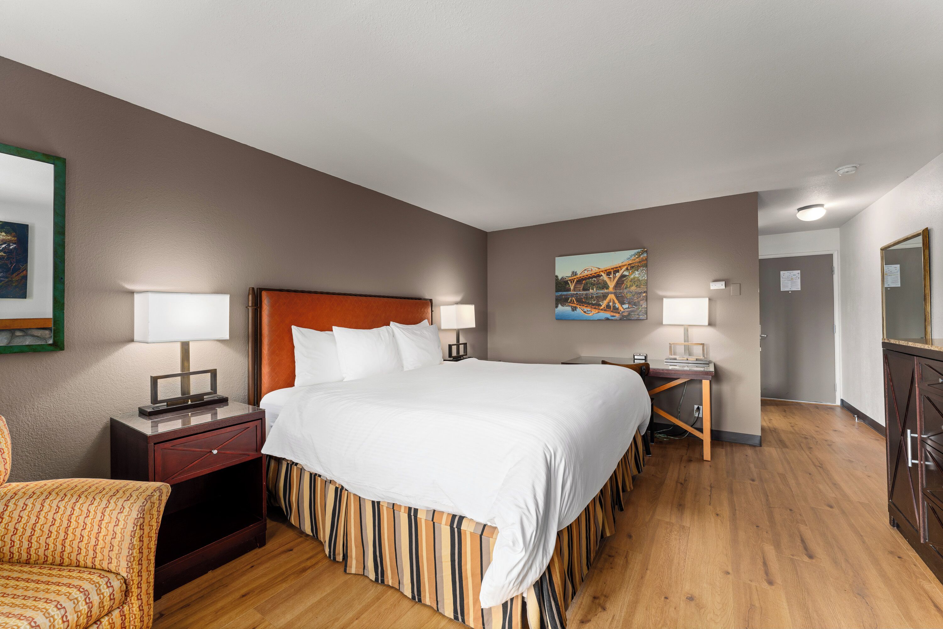 A hotel room with a bed, bedside tables, a desk, a chair, and a painting on the wall.