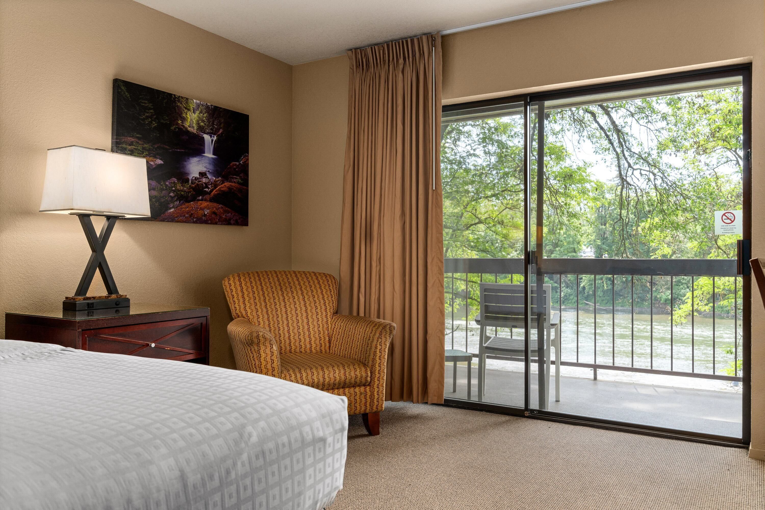 A hotel room with a bed, a chair, a painting, a lamp, and a balcony with a view of greenery and a river.