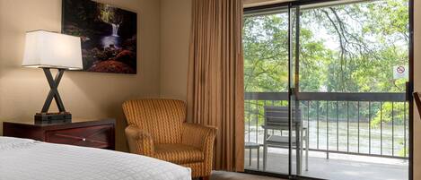 A hotel room with a bed, a chair, a painting, a lamp, and a balcony with a view of greenery and a river.