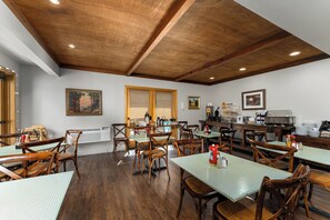 A dining area with wooden tables and chairs, a kitchen in the background, and artwork on the walls.
