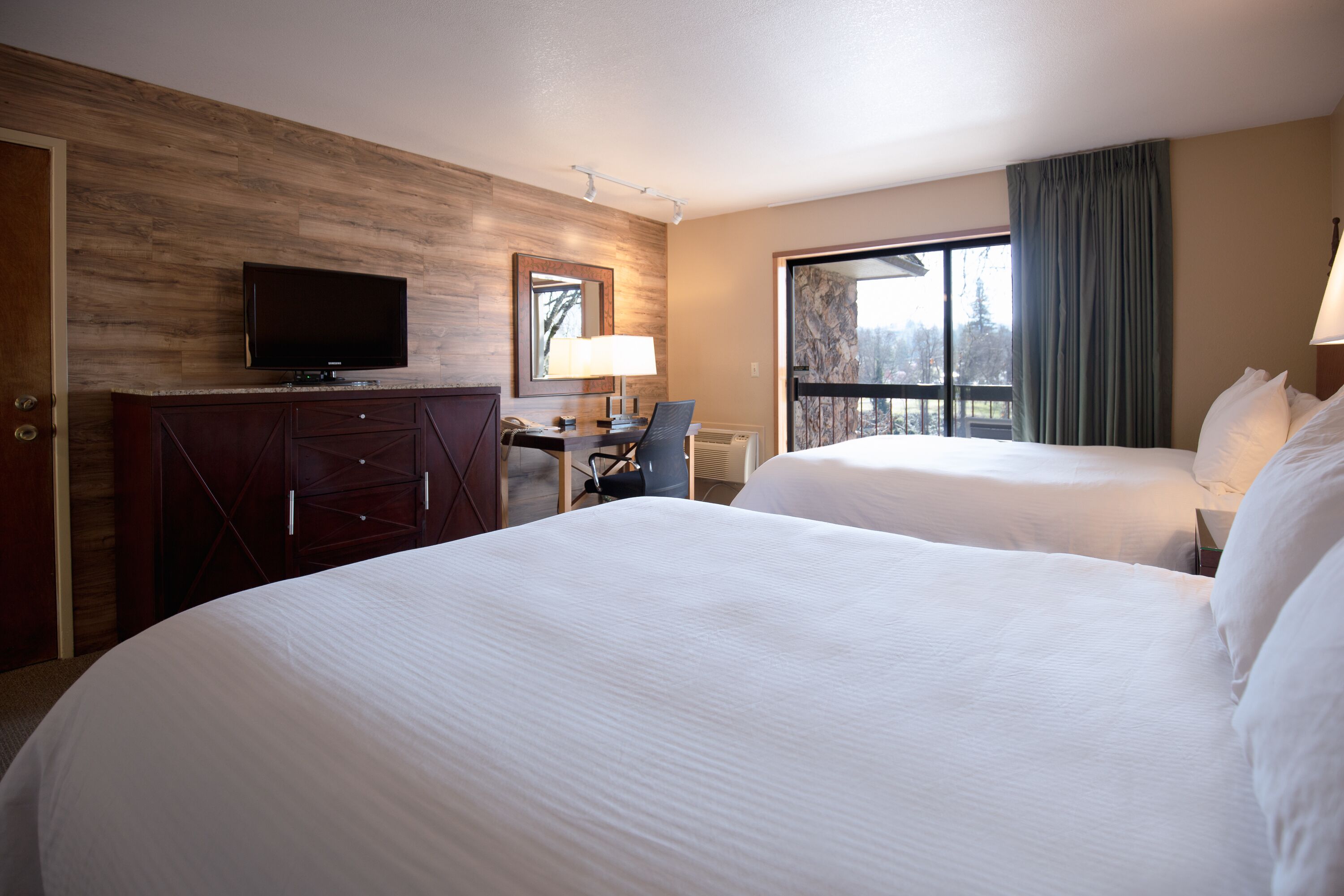Habitación de hotel con dos camas, un escritorio con silla, televisión y un ventanal con cortinas.