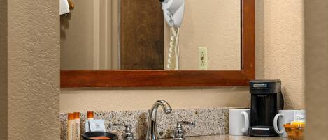 A bathroom with a sink, mirror, hair dryer, coffee maker, and toiletries.