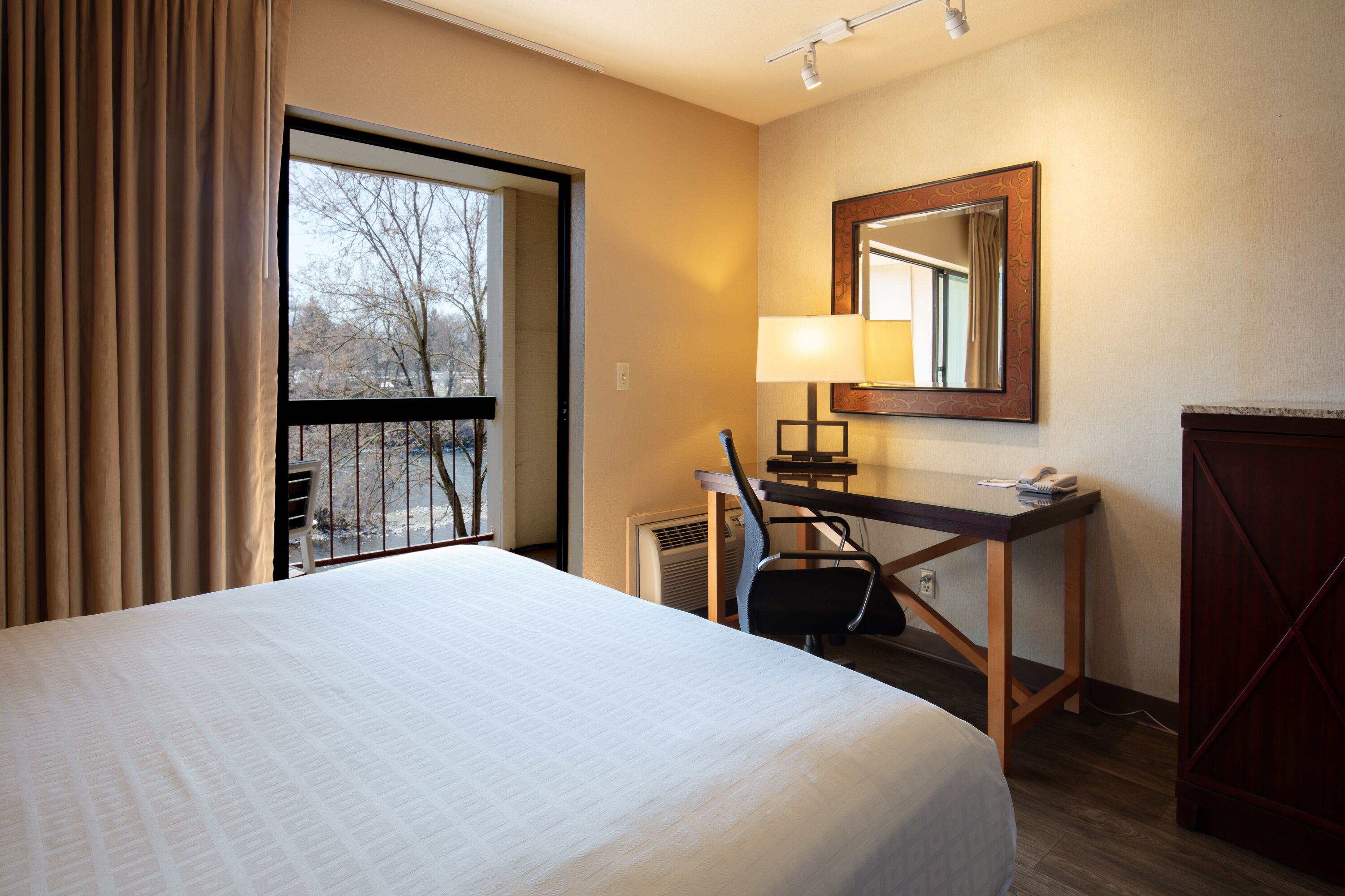 A hotel room with a large bed, a desk with a lamp, a mirror, and a view of trees through a window.