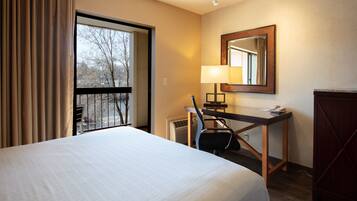 A hotel room with a large bed, a desk with a lamp, a mirror, and a view of trees through a window.