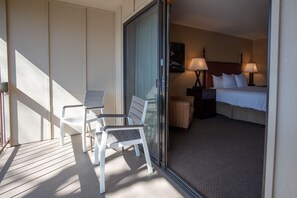 A hotel room with a bed, two bedside lamps, a chair, a small table, and a glass door leading to a balcony with a chair and a table.
