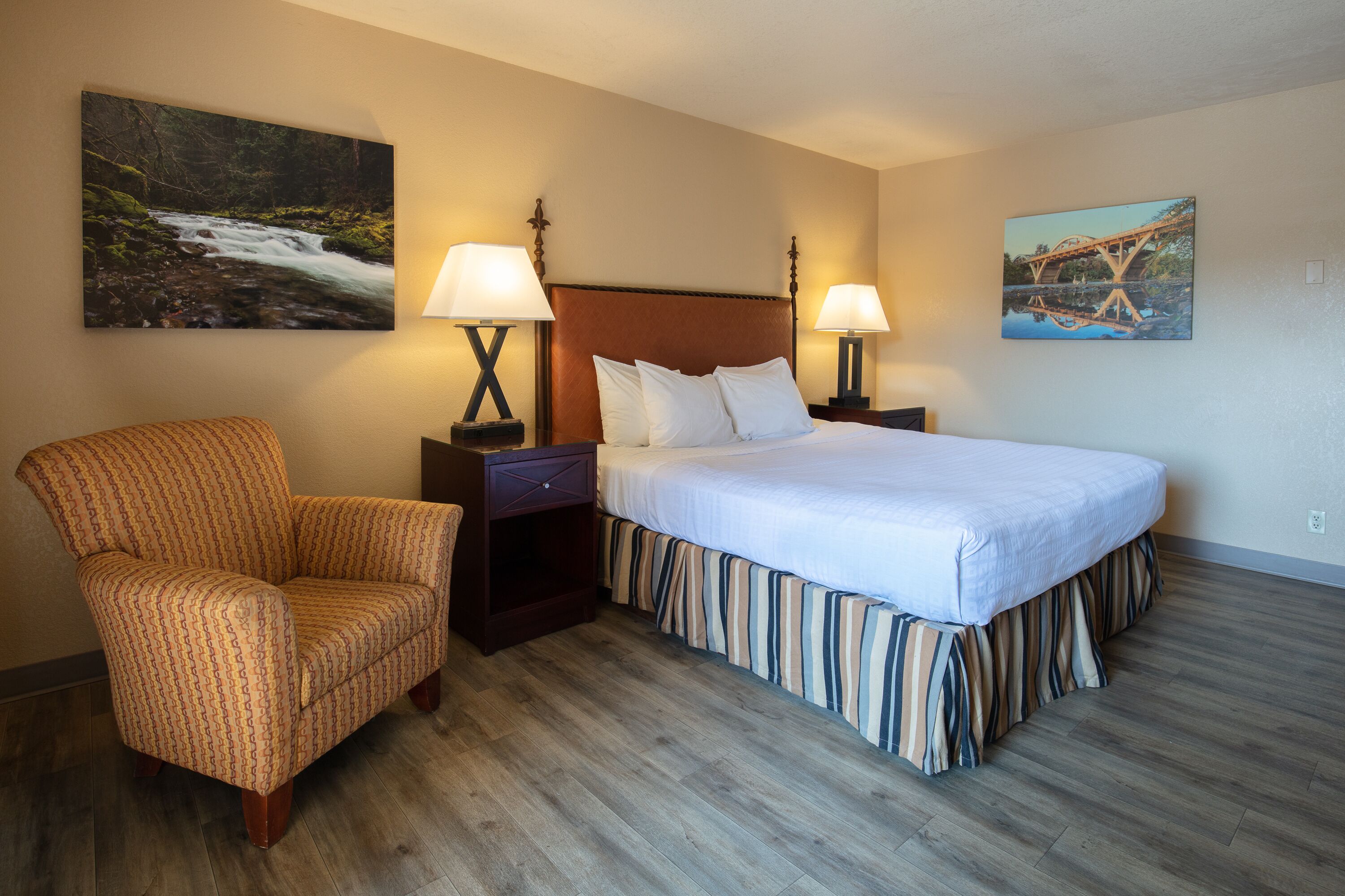 A hotel room with a bed, two bedside lamps, a chair, and two wall-mounted paintings.