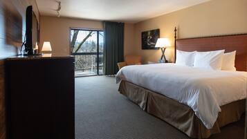 A hotel room with a large bed, a desk with a flat-screen TV, a chair, and a window with a view of trees.