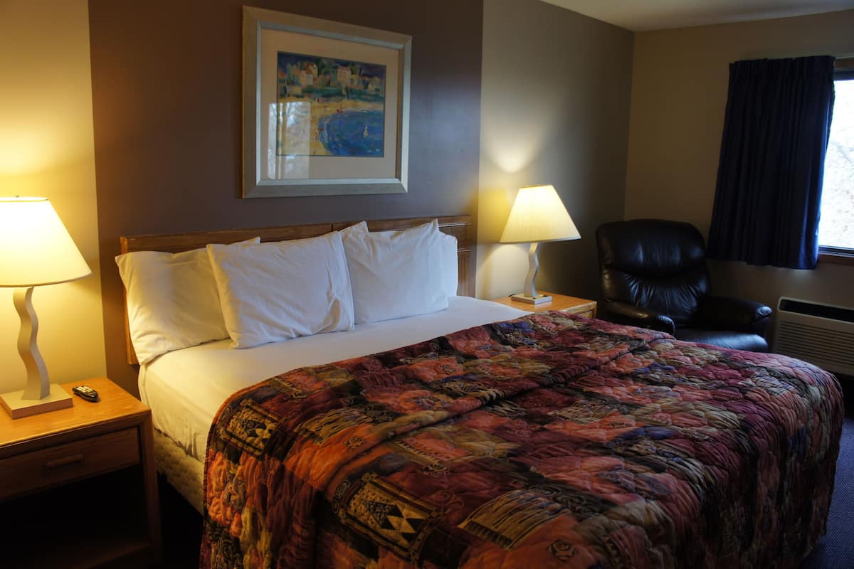 Quarto de hotel com uma cama grande, duas luminárias de cabeceira, uma cadeira e um quadro na parede.