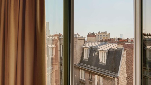 Vista a través de una ventana con cortinas, mostrando un paisaje urbano con varios edificios y tejados.