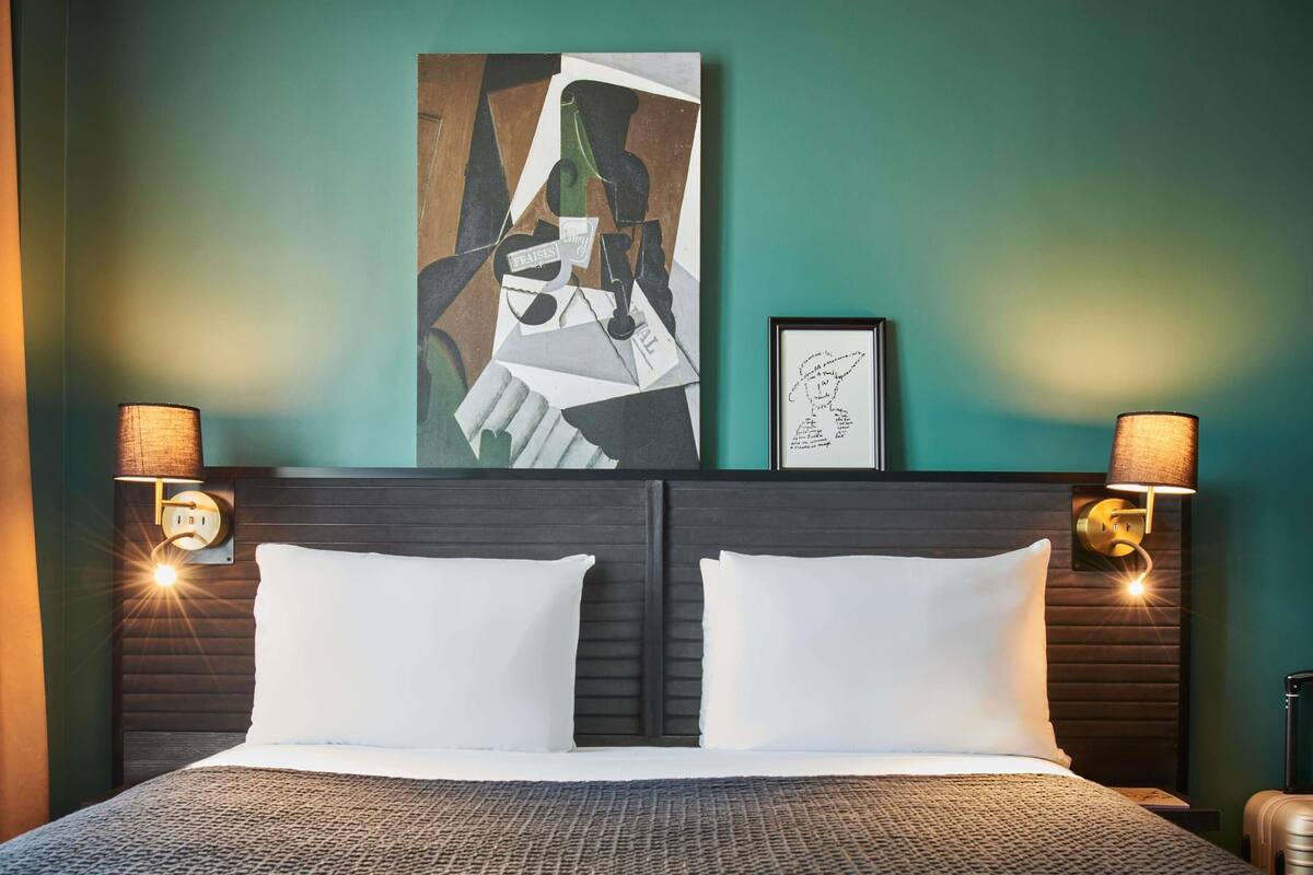 Habitación de hotel con una cama, dos lámparas fijadas a la pared, un cuadro abstracto enmarcado y una cita en la pared.