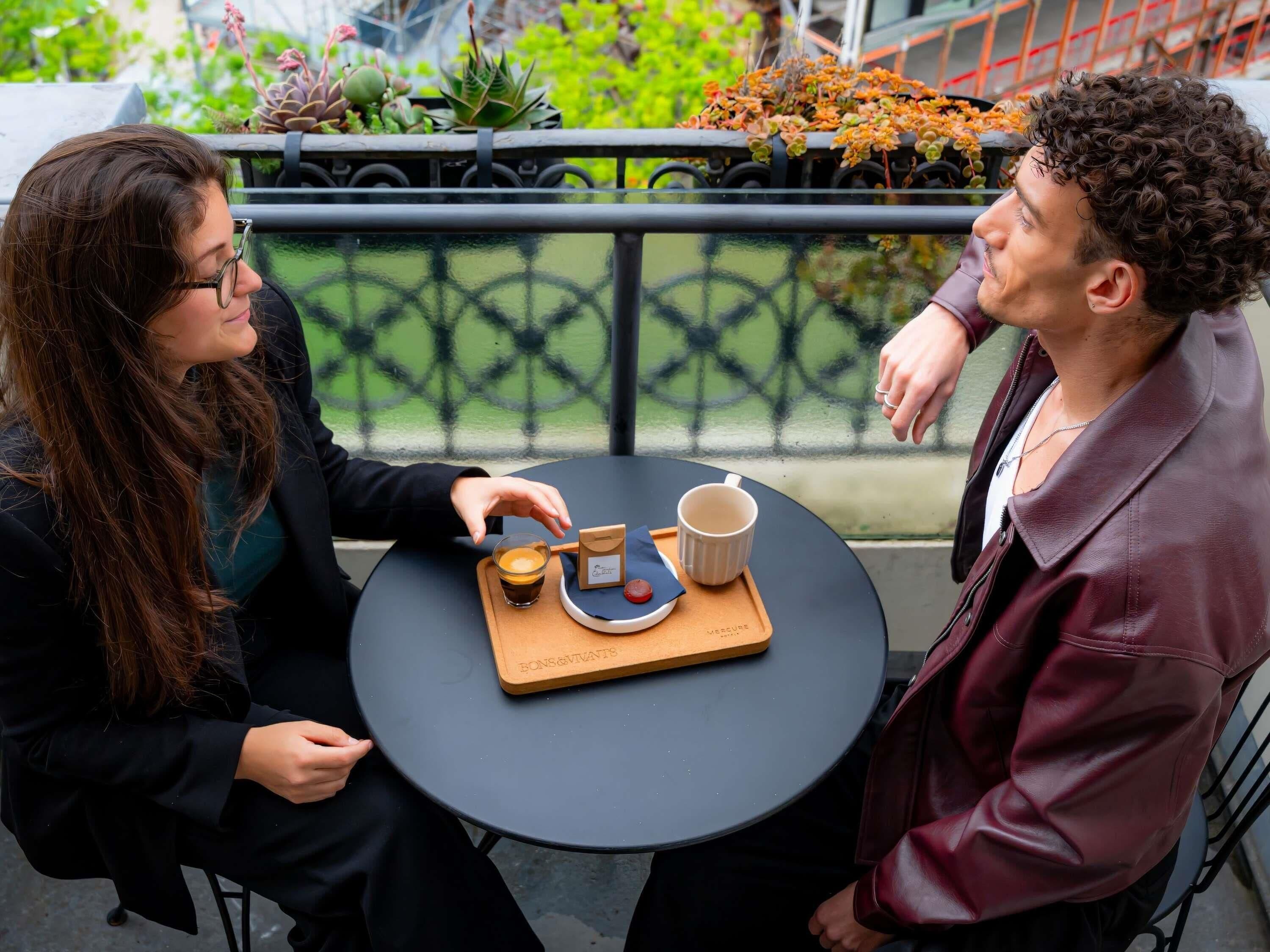 Zwei Personen sitzen an einem runden Tisch mit einem Tablett, auf dem eine Tasse und eine Schachtel stehen. Im Hintergrund sind eine Brüstung und Pflanzen zu sehen.